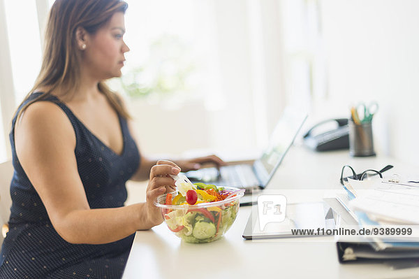 Woman eating salad and using laptop in office
