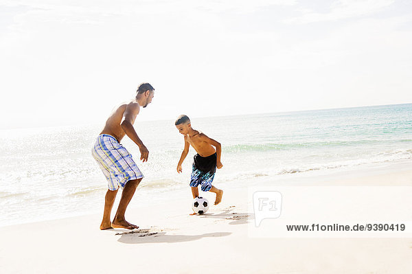 Father and son (10-11) playing soccer on beach