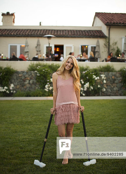 A pretty young girl stands against two croquet mallets on her two sides with her two hands rest on those and legs cross.