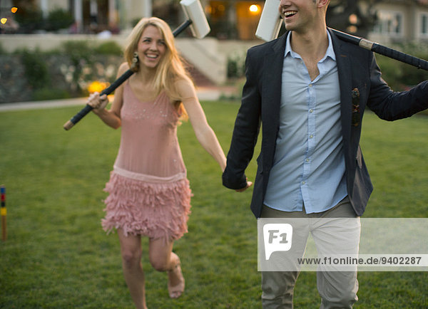 A young man with a croquet mallet on his left shoulder clutches the hand of his pretty young lady partner with a mallet on her right shoulder.