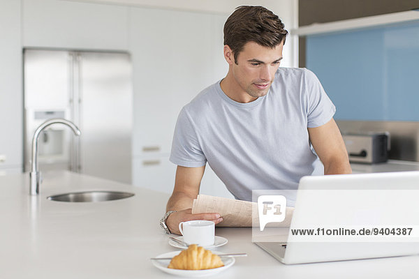 Mann mit Zeitung mit Laptop an der Küchenzeile  Kaffeetasse und Croissant im Vordergrund