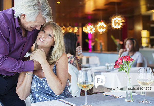 Lächelnder Mann umarmt Frau am Restauranttisch mit Weingläsern und Blumen