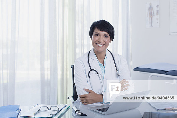 Portrait of smiling female doctor sitting with arms crossed at desk with laptop