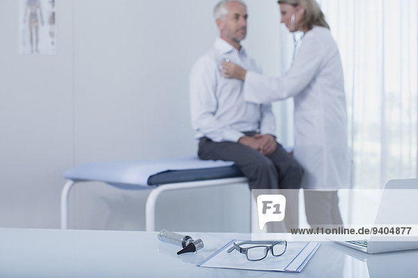 Ärztin untersucht Patientin im Büro  Laptop  Akte  Brille und Otoskop auf dem Schreibtisch im Vordergrund