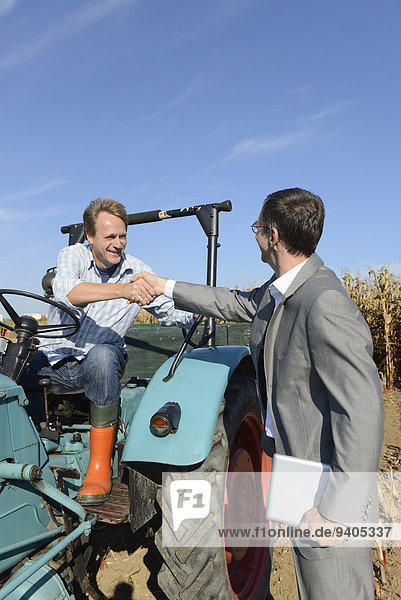 Farmer welcomes businessman with handshake while sitting on a tractor in cornfield