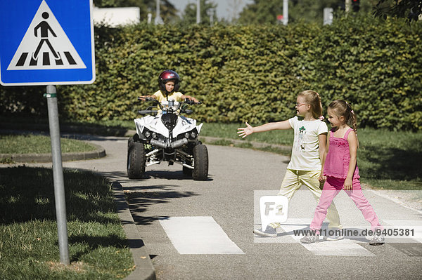 Two girls walking on zebra crossing with boy on quadbike waiting