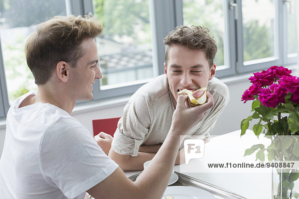 Young man feeding bread to another man  smiling