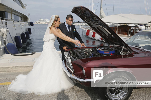 Bride and groom looking into the engine compartment of a Ford Mustang after a breakdown  at a harbour