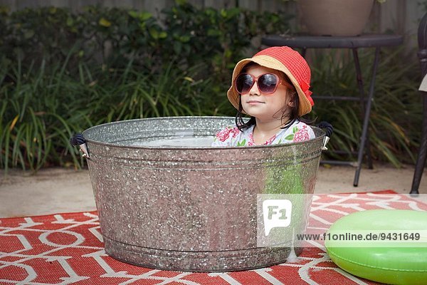 Portrait of girl wearing sunhat and sunglasses sitting in bubble bath in garden