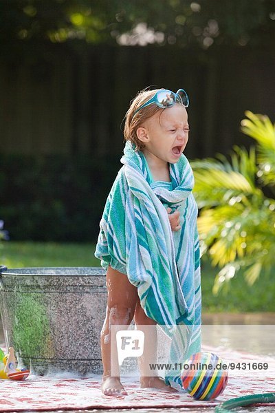 Female toddler wrapped in towel crying beside bubble bath in garden