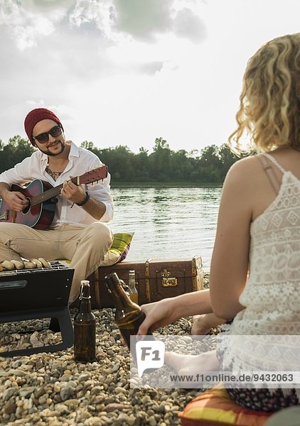 Young man sitting by lake playing guitar