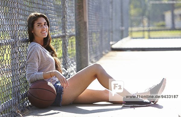 Young woman with basketball sitting by wire fence