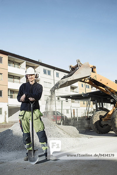 Ganzkörperporträt eines selbstbewussten Bauarbeiters mit vor Ort stehender Schaufel