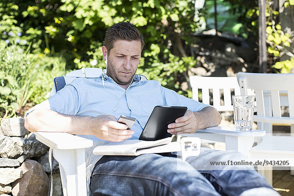 Mittlerer Erwachsener Mann mit digitalem Tablett beim Entspannen auf dem Liegestuhl im Hof