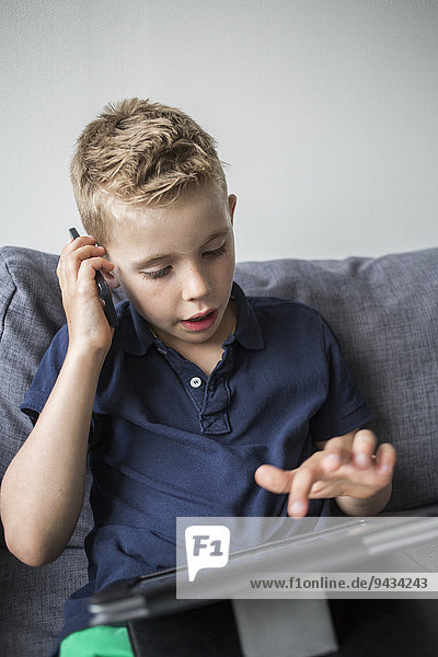 Junge beantwortet Smartphone  während er zu Hause ein digitales Tablett auf dem Sofa benutzt.