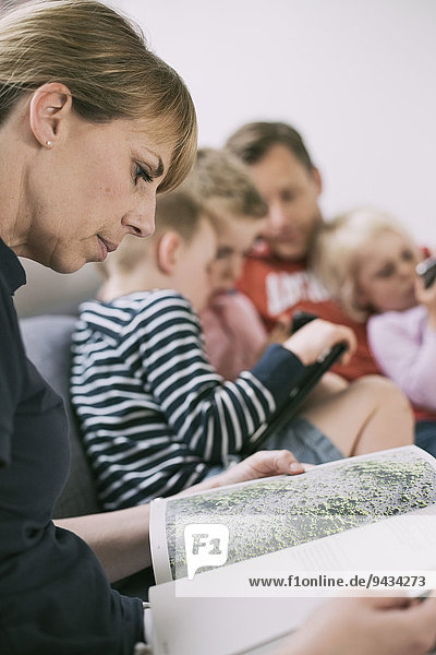 Seitenansicht des Frauenlesemagazins mit Familie unter Verwendung von Technologien im Hintergrund