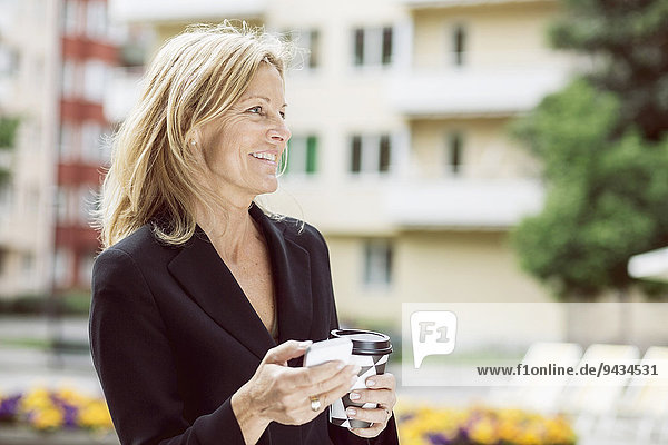Glückliche Geschäftsfrau schaut weg  während sie Smartphone und Einweg-Kaffeetasse im Freien hält.
