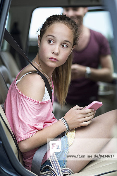 Mädchen mit Handy schaut weg  während sie im Auto sitzt  mit Vater im Hintergrund
