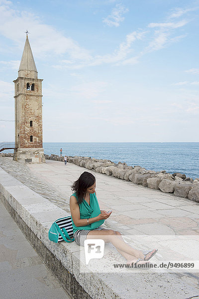 Frau mit Handy sitzt auf der Mauer vor der Kirche  Madonna dell' Angelo  Adria  Caorle  Region Venetien  Italien  Europa