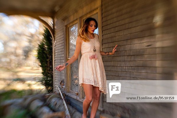 30 year old brunette woman wearing a dress balancing on some old wood on the porch of an old country house.