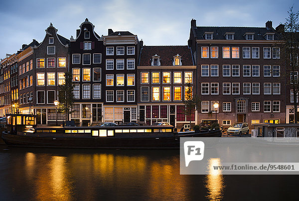 Niederlande  Amsterdam  Blick auf die Reihe der beleuchteten alten Wohnhäuser am Stadtkanal bei Abenddämmerung