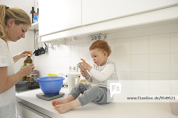 Kleines Mädchen sitzt auf der Küchentheke mit Smartphone  während ihre Mutter backt