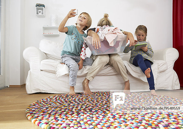 Erschöpfte Mutter mit Wäschekorb auf der Couch mit Kindern mit digitalem Tablett und Handy