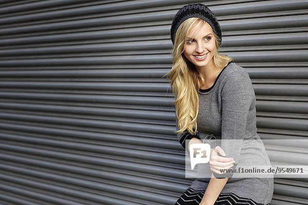 Smiling blond woman wearing knit dress and comforter standing in front of roller shutter