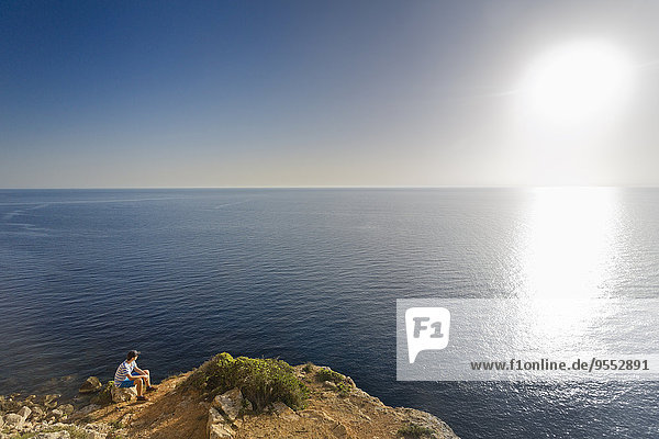 Spanien,  Balearen,  Mallorca,  ein Teenager sitzt auf einem Felsen an der Steilküste und schaut dem Sonnenuntergang zu.