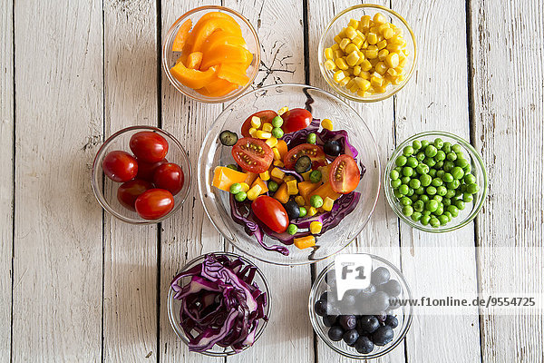 Gemischte Salat- und Glasschalen mit verschiedenen Rohkost-Regenbogenfarben auf weißem Holz arrangiert