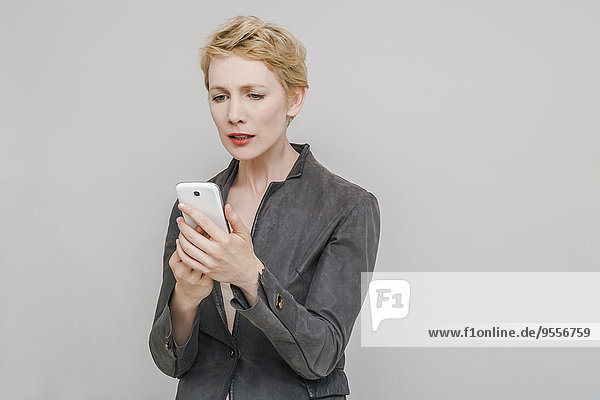 Porträt einer verstörten blonden Frau  die ihr Smartphone vor grauem Hintergrund anschaut