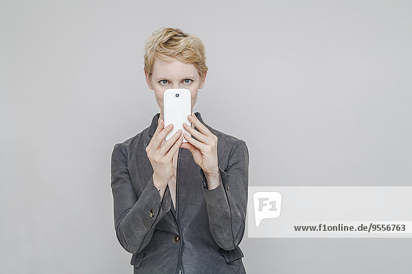 Blond woman taking a photography with smartphone in front of grey background