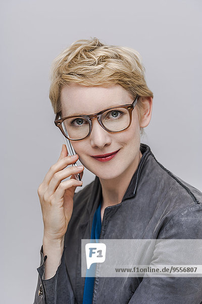 Porträt einer lächelnden  blonden Frau mit Brille  die vor grauem Hintergrund telefoniert.