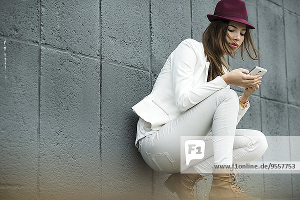 Junge Frau mit Hut blickt auf ihr Smartphone vor der grauen Wand