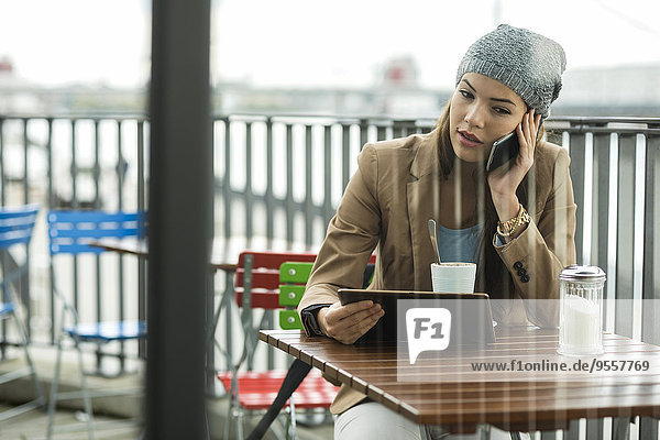 Junge Frau sitzt auf dem Bürgersteig und hält ein digitales Tablett beim Telefonieren mit dem Smartphone.