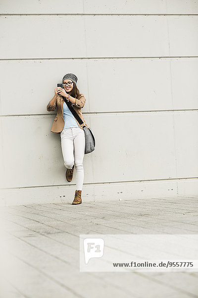 Junge Frau,  die sich an eine Wand lehnt und einen Selfie mit ihrem Smartphone nimmt.