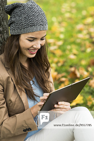 Lächelnde junge Frau  die sich mit einem digitalen Tablett an einen Baum lehnt.