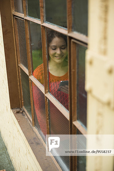 Lächelnde junge Frau mit Handy hinter dem Fenster