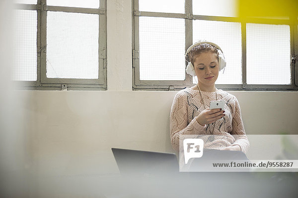 Junge Frau auf dem Schreibtisch sitzend mit Kopfhörer und Handy