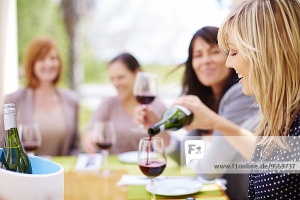 Frau  die mit Freunden im Hintergrund in ein Glas Rotwein gießt.