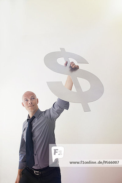 Zuversichtlicher Geschäftsmann hält Dollarzeichen gegen die Wand im Büro hoch