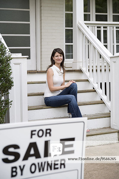 Eine junge Frau sitzt auf der Treppe eines Hauses hinter einem Schild Zu verkaufen durch den Besitzer .