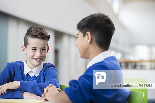 Two smiling schoolboys wearing school uniforms talking at desk in classroom