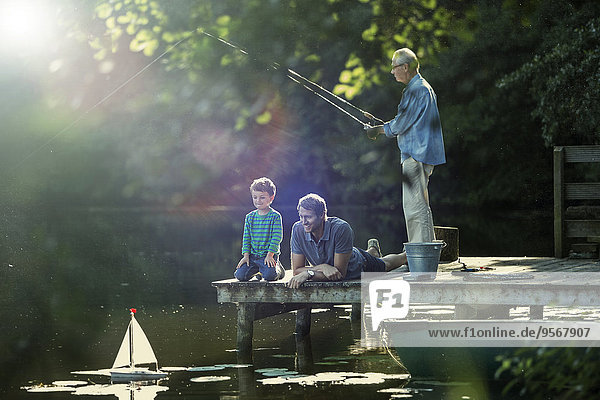 Junge fischen und spielen mit Spielzeug-Segelboot mit Vater und Großvater am See