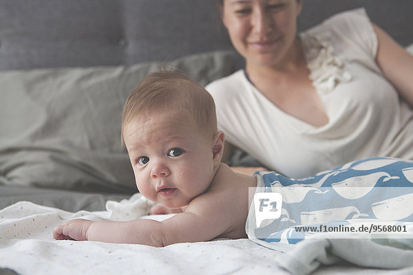 Porträt des kleinen Babys auf dem Bett liegend mit lächelnder Mutter im Hintergrund