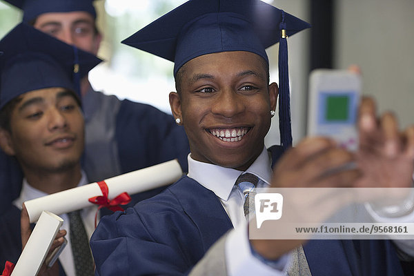 Smiling male student wearing graduation clothes taking selfie with friends