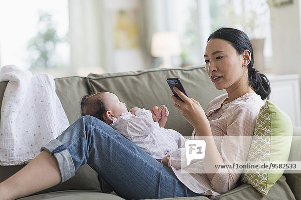 Asian mother using cell phone and holding baby in living room