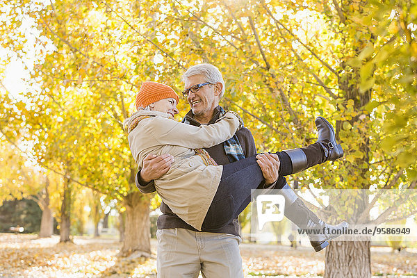 Older Caucasian man carrying wife under autumn trees