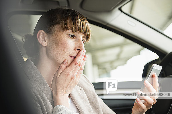 Porträt einer Geschäftsfrau  die in einem Auto sitzt und ihr Smartphone mit der Hand auf dem Mund betrachtet.