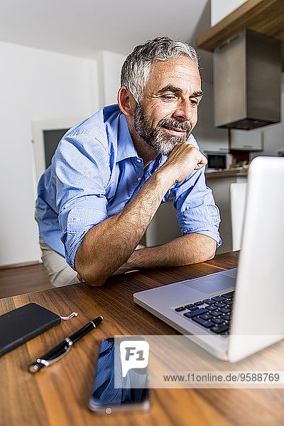 Porträt eines lächelnden Geschäftsmannes im Home-Office mit Blick auf seinen Laptop